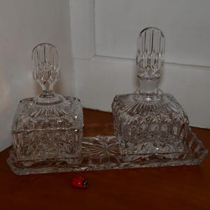 Könnte beinhalten: Zwei durchsichtige Glasflaschen mit Stopfen, die auf einem durchsichtigen Glastablett stehen. Die Flaschen haben ein rautenförmiges Muster und stehen auf einer Holzoberfläche.