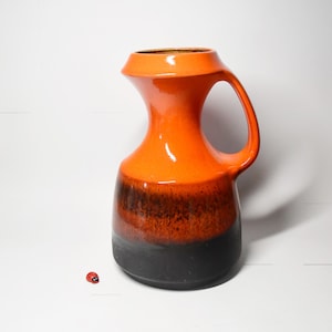Può includere: Una brocca in ceramica con manico, caratterizzata da una sfumatura di colori. La parte superiore è arancione brillante, che sfuma in una fascia marrone scuro e termina con una base nera. Una piccola statuina di coccinella è accanto alla brocca.