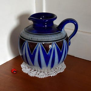 Puede incluir: Una jarra de cerámica azul cobalto con asa y boquilla. La jarra presenta un patrón decorativo de zigzags blancos y azules y una banda marrón. Se encuentra sobre un tapete de encaje blanco. Una pequeña mariquita roja está al lado del tapete.