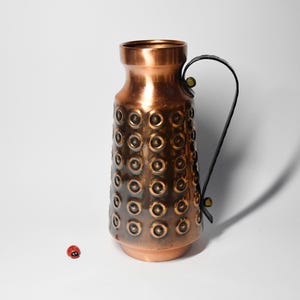 Può includere: Un vaso di rame con un motivo circolare testurizzato e un manico in pelle nera. Il vaso è su una superficie bianca.