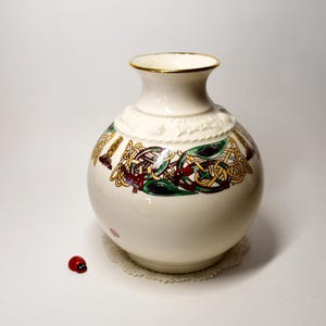 May include: A cream-coloured ceramic vase with a round body and a narrow neck, decorated with Celtic knot designs in green, gold, and red. The rim is trimmed with gold. The vase sits on a white lace doily.