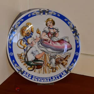 Könnte beinhalten: Ein weißer Keramikteller mit blauem Rand und einer bunten Illustration einer Frau in Tracht, einem Mann und einem Löwen. Der Teller trägt den Text "Das Schuhplattln" in blauer Schrift.