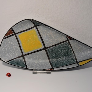 Puede incluir: Un plato de servir de cerámica con un patrón geométrico en tonos de gris, verde, amarillo y marrón. El plato tiene forma de media luna y tiene un esmalte moteado.