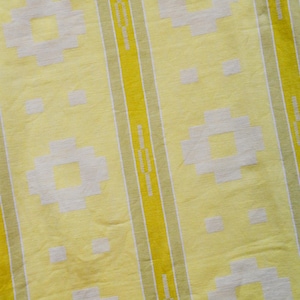 Könnte beinhalten: Nahaufnahme eines gelben Stoffes mit einem sich wiederholenden Muster. Das Design zeigt vertikale Streifen in Gelb und Hellgrün mit weißen geometrischen Formen und Quadraten. Der Stoff hat eine leicht zerknitterte Textur.