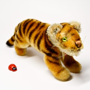 Può includere: Un giocattolo di tigre di peluche con strisce arancioni e nere, occhi verdi e un naso rosa. Un piccolo giocattolo di coccinella rossa è di fronte alla tigre. La tigre ha un aspetto vintage.