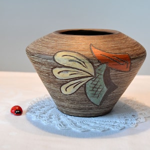 Puede incluir: Un jarrón de cerámica marrón con un diseño estilizado de un pájaro en amarillo, verde y naranja. El jarrón tiene una boca ancha y una base estrecha.