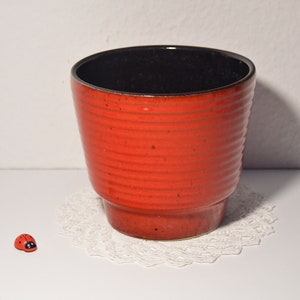 Könnte beinhalten: Ein roter Keramiktopf mit schwarzem glasiertem Innenraum. Der Topf hat ein geripptes Design und eine leicht ausgestellte Lippe.