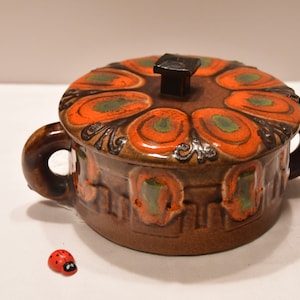 Puede incluir: Un plato cubierto de cerámica marrón con diseños florales naranja y verde. La tapa tiene un pomo cuadrado.