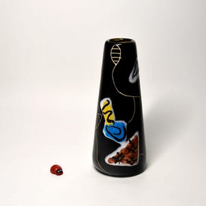 Könnte beinhalten: Eine schwarze Vase mit abstrakten, farbenfrohen Designs und goldenen Akzenten. Die Vase hat einen schmalen Hals und einen breiteren Boden. Eine kleine rote Marienkäfer-Figur liegt neben der Vase. Die Vase wird wahrscheinlich zu Dekorationszwecken verwendet.