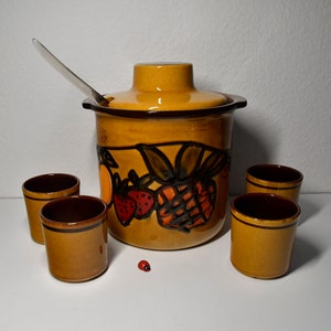 Könnte beinhalten: Ein Set aus sechs Keramikbechern und einem großen Keramik-Krug mit Deckel. Der Krug hat einen braunen Rand und ein buntes Design mit Früchten, darunter Erdbeeren, Ananas und Orangen. Die Becher sind braun mit einem gelben Rand.