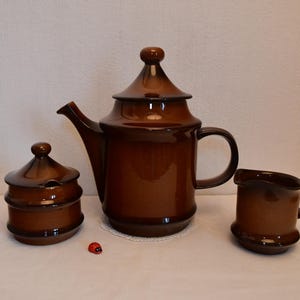 Peut inclure: Un service à thé en céramique marron, comprenant une théière avec un couvercle, un sucrier avec un couvercle et une crémière. L'ensemble a une finition brillante et un design classique. Une figurine de coccinelle rouge est également présente.