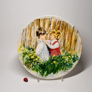 Könnte beinhalten: Ein dekorativer Keramikteller mit einer gemalten Szene von zwei Kindern in einem Garten. Der Teller hat einen Holzzaunhintergrund und ist von Blumen umgeben. Eine kleine Marienkäferfigur befindet sich vor dem Teller.