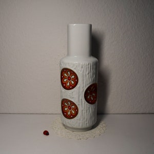 Könnte beinhalten: Weiße Keramikvase mit strukturierter Oberfläche und drei orangefarbenen und weißen Blumendesigns.