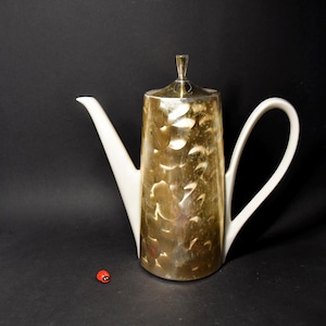 Könnte beinhalten: Eine weiße Kaffeekanne aus Keramik mit goldenem Metallic-Design. Die Kanne hat einen goldenen Deckel und einen weißen Henkel.