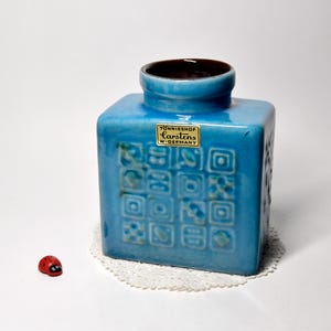 May include: A turquoise ceramic vase with a square shape and a dark brown interior. The vase features a decorative pattern and a label reading "Tonnieshof Carstens W-Germany". A small red ladybird figurine is also present.