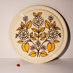 Könnte beinhalten: Runder, cremefarbener Keramik-Tablett mit floralem Design. Das Design zeigt braune Stiele, gelbe Blätter und weiße und gelbe Blüten. Vor dem Tablett sitzt ein kleiner roter Marienkäfer.