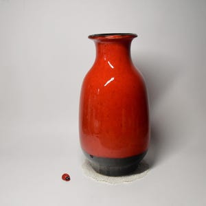 Może przedstawiać: Wysoki, błyszczący ceramiczny wazon z żywą czerwoną górną częścią i ciemnoszarą podstawą. Wazon ma zaokrąglony korpus i wąską szyjkę. W pobliżu stoi mała figurka biedronki. Wazon ma około 25 cm wysokości.