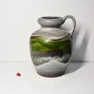 Può includere: Un vaso in ceramica con manico, nei toni del verde, grigio e marrone. Il vaso ha un corpo bulboso e un collo stretto. Il design include una fascia verde e una finitura strutturata e macchiettata. Una piccola coccinella rossa è sulla superficie.