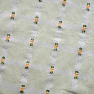 Könnte beinhalten: Nahaufnahme eines hellgrünen Stoffes mit einem Gittermuster. Vertikale weiße Streifen kreuzen sich mit horizontalen weißen Streifen. Die Kreuzungen sind mit kleinen Clustern aus gelben und grünen Stickereidetails verziert.