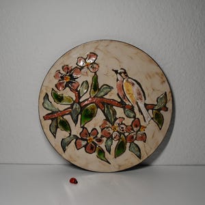 Könnte beinhalten: Ein weißer Keramikteller mit einem erhabenen Design eines Vogels, der auf einem Zweig mit rosa und grünen Blumen sitzt. Der Teller hat ein rustikales, vintage-mäßiges Aussehen.