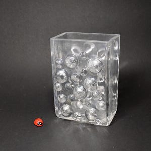 Può includere: Vaso rettangolare in vetro trasparente con un design a bolle. Il vaso ha una forma quadrata ed è decorato con bolle circolari in rilievo di varie dimensioni. Accanto al vaso c'è una piccola statuina di coccinella rossa.