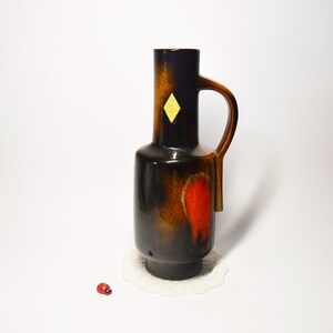 Könnte beinhalten: Eine Vintage-Keramikvase mit Henkel, mit dunkelbrauner und orangefarbener Glasur und rotem Akzent. Die Vase hat einen gelben rautenförmigen Aufkleber. Sie steht auf einer weißen Spitze, mit einer kleinen Marienkäferfigur in der Nähe.