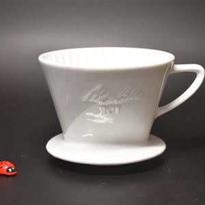 May include: A white ceramic coffee dripper with a handle and a circular base. The side of the dripper features embossed text that reads Melitta 101. A small red and black ladybird figurine is positioned to the left of the coffee dripper.