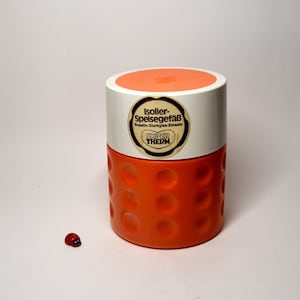 Könnte beinhalten: Ein orangefarbener und weißer isolierter Lebensmittelbehälter mit runder Form und Deckel. Der Behälter hat ein strukturiertes Muster aus kreisförmigen Vertiefungen. Das Etikett lautet "Isolier-Speisegefäß" und "SUPER THERM". Eine kleine Marienkäferfigur befindet sich im Vordergrund.