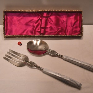 Könnte beinhalten: Ein silbernes Gabel- und Löffelset mit weißen marmorierten Griffen in einer mit rosa Satin ausgekleideten Schachtel.