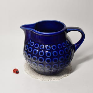 Op de afbeelding: Een kobaltblauwe keramische kan met een gestructureerd, gestippeld patroon. De kan heeft een handvat en een tuit. Een klein rood lieveheersbeestje beeldje staat ernaast op een wit oppervlak.
