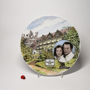 Può includere: Piatto decorativo in ceramica con un'illustrazione a colori di un edificio con tetto rosso, circondato da alberi e un sentiero. Un ritratto di un uomo e una donna è a destra. Il piatto è su un supporto trasparente.