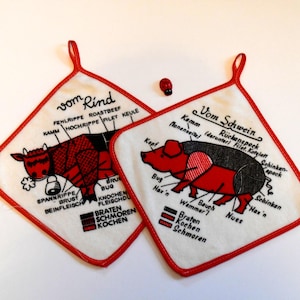 Könnte beinhalten: Zwei weiße Topflappen mit rotem Rand und einer schwarz-roten Illustration einer Kuh und eines Schweins. Der Topflappen mit der Kuh hat den Text "vom Rind" und der Topflappen mit dem Schwein hat den Text "Vom Schwein".