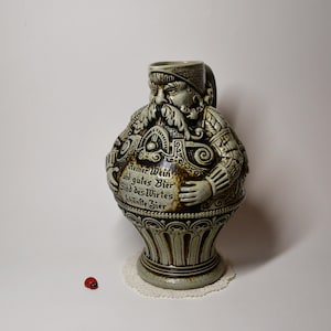 Peut inclure: Une chope en céramique en forme d'homme trapu avec une longue barbe, portant un chapeau et se tenant le ventre. La chope est grise et marron avec des détails et du texte ornés. Une petite coccinelle rouge est à proximité.