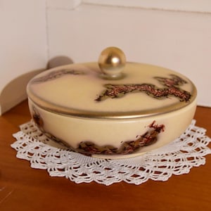 Peut inclure: Une boîte à bijoux en céramique blanche avec un couvercle orné d'un bord doré et d'un motif en spirale brun et or. La boîte est posée sur un napperon en crochet blanc.