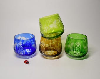 May include: Four glass tumblers in blue, light green, amber, and dark green, each featuring intricate etched floral and geometric patterns. A small red ladybug figurine is positioned on the surface near the blue glass tumbler.