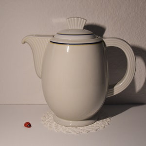 Könnte beinhalten: Weiße Kaffeekanne aus Keramik mit blauem und goldenem Rand und einem Deckel mit einem dekorativen Knauf. Die Kanne steht auf einer weißen Spitze.