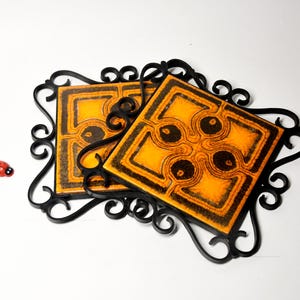 Könnte beinhalten: Zwei quadratische dekorative Fliesen mit einem orange-schwarzen Muster, eingefasst in einem schwarzen Schmiedeeisenrahmen. Ein kleiner roter Marienkäfer befindet sich links von den Fliesen. Die Fliesen sind wahrscheinlich für die Heimdekoration.