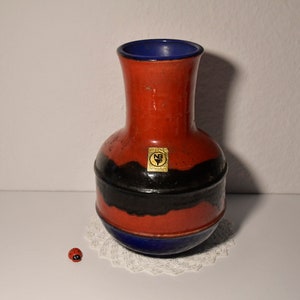 May include: A red and black ceramic vase with a blue rim and a gold label that reads "W.R. Röschlau" and has a logo with the letters "NB". The vase is sitting on a white doily.
