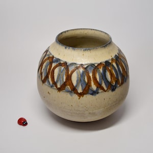 Può includere: Un vaso in ceramica rotondo color crema con una consistenza maculata. La parte superiore presenta un motivo ripetitivo di forme ovali dipinte a mano in blu e marrone che circondano il collo. Una piccola statuetta di coccinella rossa e nera è posizionata accanto.
