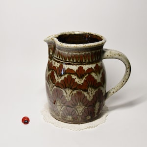 Könnte beinhalten: Keramikkrug mit Henkel und Ausguss, verziert mit einem braunen und cremefarbenen Blumenmuster. Der Krug steht auf einer weißen Spitze, mit einem kleinen roten Marienkäfer daneben. Die Keramik hat ein rustikales Aussehen.