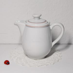 Könnte beinhalten: Eine weiße Keramik-Teekanne mit Deckel, Ausguss und Henkel. Der Deckel hat einen dünnen roten und blauen Streifen. Die Teekanne steht auf einer weißen Spitzendeckchen. Eine kleine rote Marienkäfer-Figur befindet sich links vom Deckchen.