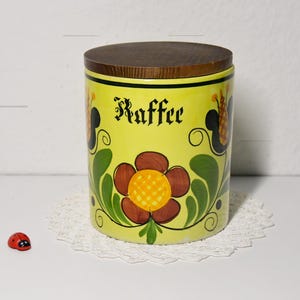 Può includere: Un barattolo di caffè cilindrico in ceramica gialla con coperchio in legno. La parola "Kaffee" è scritta in nero. Il barattolo è decorato con fiori marroni e gialli e foglie verdi. È visibile una piccola statuina di coccinella rossa.