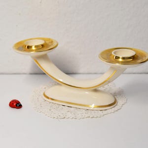 Könnte beinhalten: Cremefarbener Kerzenhalter mit zwei Armen und goldenen Verzierungen. Die geschwungenen Arme tragen zwei Kerzenschalen mit goldenen Rändern. Ein kleiner roter Marienkäfer befindet sich in der Nähe. Der Kerzenhalter steht auf einer weißen Spitze.