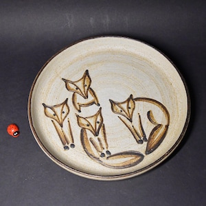 Op de afbeelding: Een crèmekleurige keramische plaat met een bruine rand en een grillig vossendesign. Vier gestileerde vossen zijn afgebeeld in bruin, in verschillende poses. Een kleine rode lieveheersbeestje is zichtbaar links van de plaat.