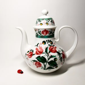 Könnte beinhalten: Weiße Keramik-Teekanne mit Deckel, verziert mit roten Granatapfel- und grünen Blattmustern. Die Teekanne hat einen gebogenen Ausguss und Henkel. Eine kleine rote Marienkäfer-Figur steht daneben. Küchenartikel.