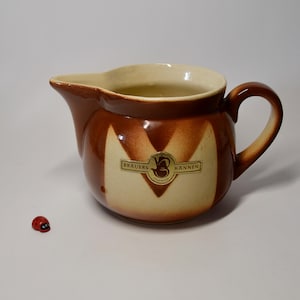 Op de afbeelding: Een bruine en crèmekleurige keramische kan met een handvat. De kan heeft een bruin en crèmekleurig gestreept patroon en een logo met de tekst "Brauers Kannen".