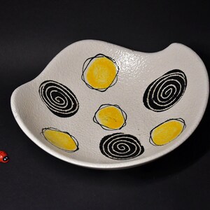 Può includere: Una ciotola in ceramica bianca con bordo ondulato, decorata con disegni a spirale gialli e neri. La ciotola ha una superficie strutturata ed è posta su uno sfondo nero. Una piccola coccinella rossa si trova nell'angolo in basso a sinistra.