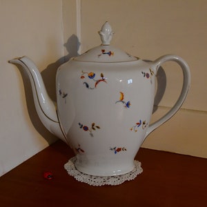 Könnte beinhalten: Weiße Porzellan-Teekanne mit einem Blumenmuster in Blau-, Orange- und Gelbtönen. Die Teekanne hat einen Deckel mit einem Knopf in Form einer Blume. Die Teekanne steht auf einer weißen Spitzenunterlage.