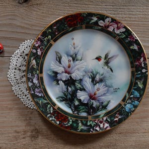 Könnte beinhalten: Dekorative Platte mit Blumen- und Kolibri-Design. Die Platte zeigt ein zentrales Bild von weißen Hibiskusblüten und einem Kolibri, umgeben von einem Rand aus Blumen und grünen Blättern. Die Platte hat einen goldenen Rand.