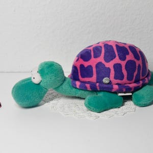 Può includere: Una tartaruga di peluche con un guscio rosa e viola e una testa e un corpo verdi. La tartaruga è sdraiata a pancia in giù con la testa tesa.